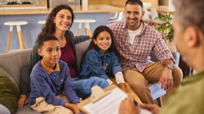 family therapy - Road to Healing Counseling Smiling family of four on a sofa in a cozy living room, listening to someone with papers in hand.
